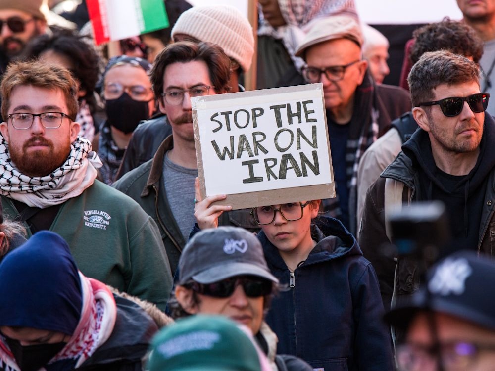 A crowd of solemn-looking people. One person holds up a sign that says &lsquo;Stop the war on Iran.&rsquo;