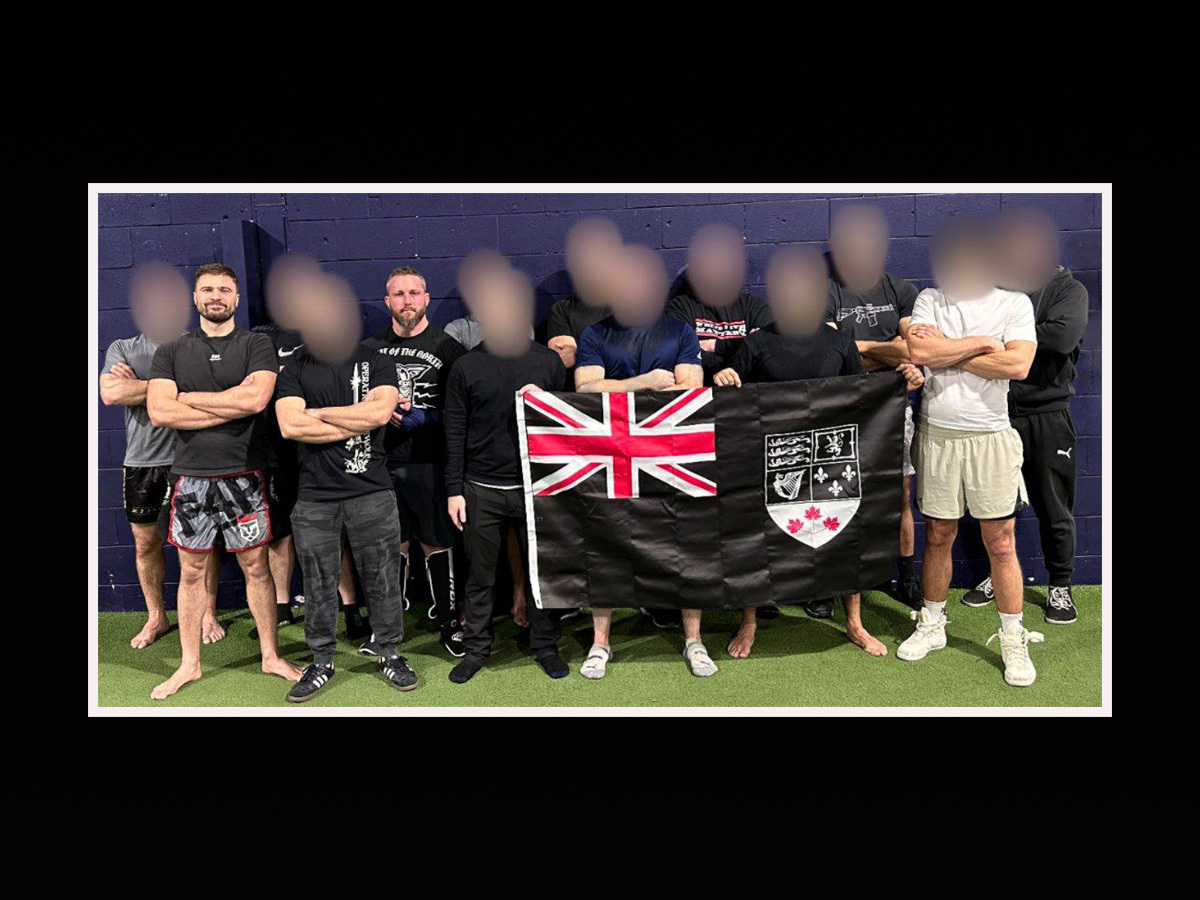 Eleven men stand in a row wearing shorts and other athletic gear. Nine of the men have their faces blurred. They hold a version of Canada&rsquo;s red ensign flag, but instead of a red background, the flag&rsquo;s background is black.