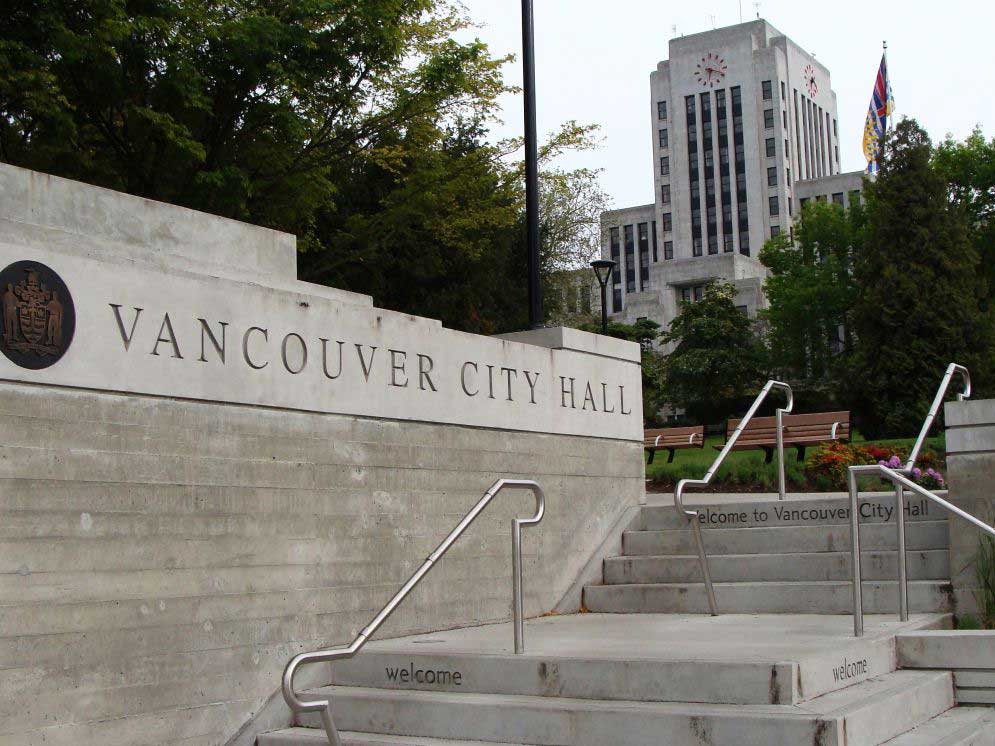 A sign saying Vancouver City Hall, and stairs with the word welcome etched into them. In the background an Art Moderne style building with a clock.