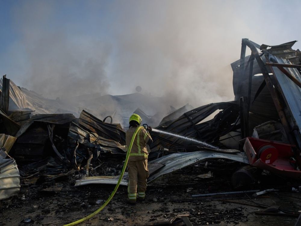 A person in mustard and flourescent protective clothing stands amidst the smoking wreckage of a building hit by a missile strike and holds on their shoulder a hose shooting water into the wreckage.