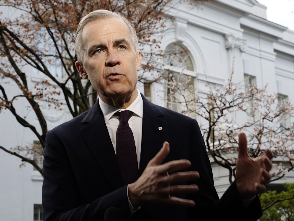 A grey-haired, white, late-middle-aged man in a suit and tie, Canada&rsquo;s prime minister, gestures with his hands in front of a white building of classical architectural design.