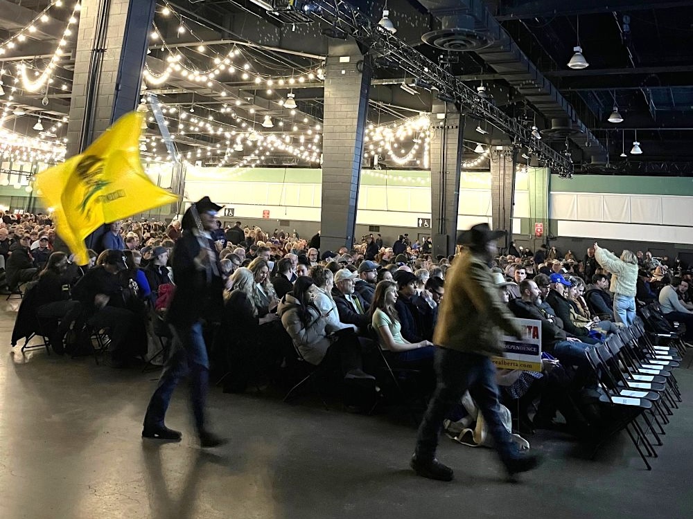 A large hall with twinkling lights overhead is filled with seated people. In the foreground two men with cowboy hats walk by, one waving a yellow flag.
