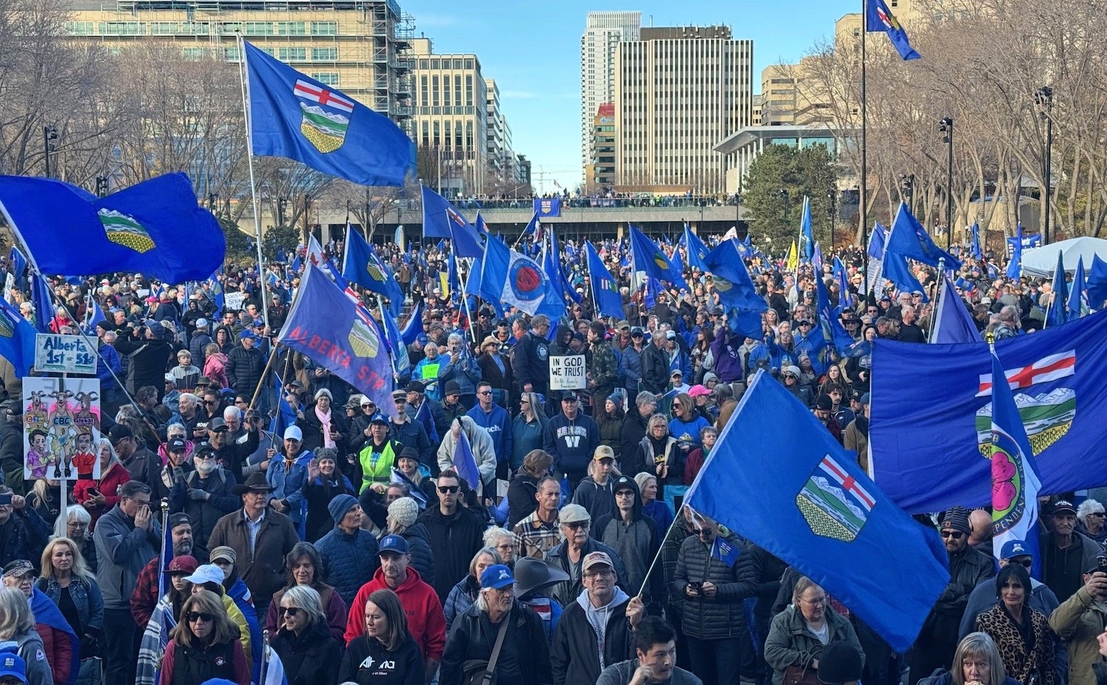 A crowd of people on a sunny day holding flags that symbolize an independent Alberta and placards with slogans including &lsquo;Alberta First.&rsquo;