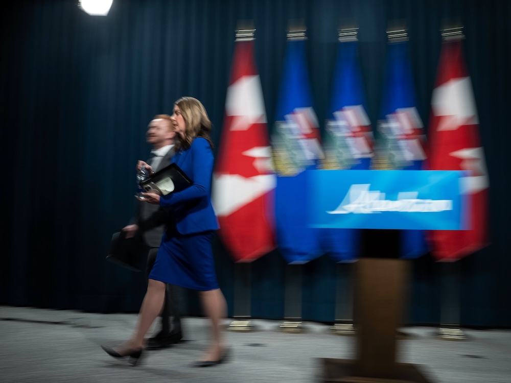 Alberta Premier Danielle Smith has shoulder-length brown hair. She is wearing a blue suit and walking offstage towards the left of the frame with a man in a dark suit and white shirt to her right side against a wall of dark blue curtains with a standing row of flags and a blurry wooden podium with a blue sign on it.