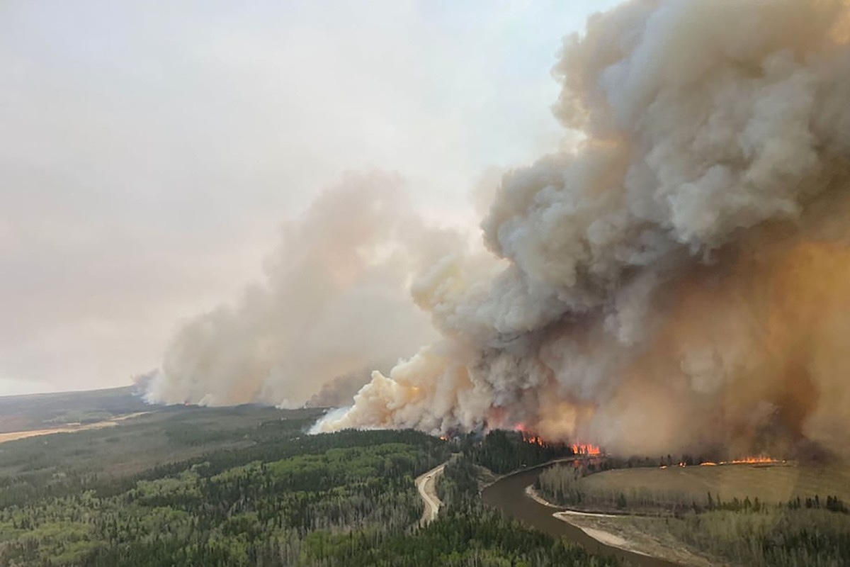 A large wildfire emits billowing clouds of smoke as it burns across a flat forest. Flames are seen near a river.