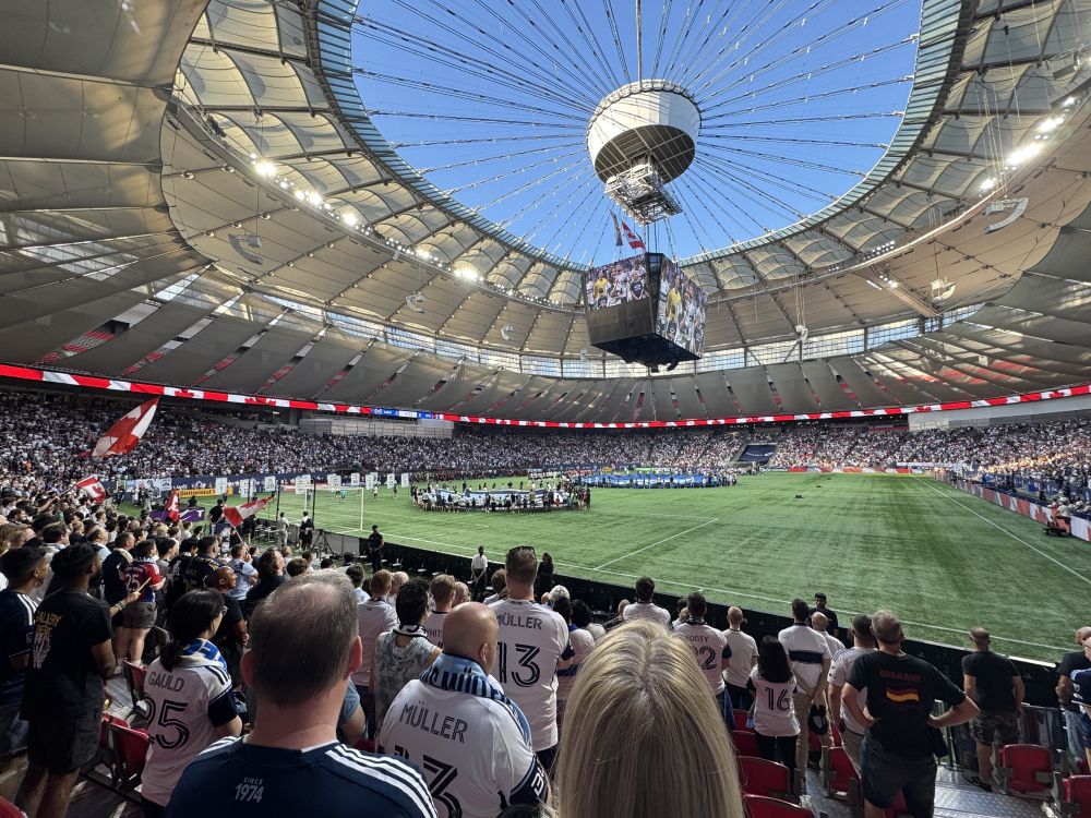 Crowds of people in sports jerseys stand in the seating areas of an open-air stadium and watch the opening ceremony of a soccer match.