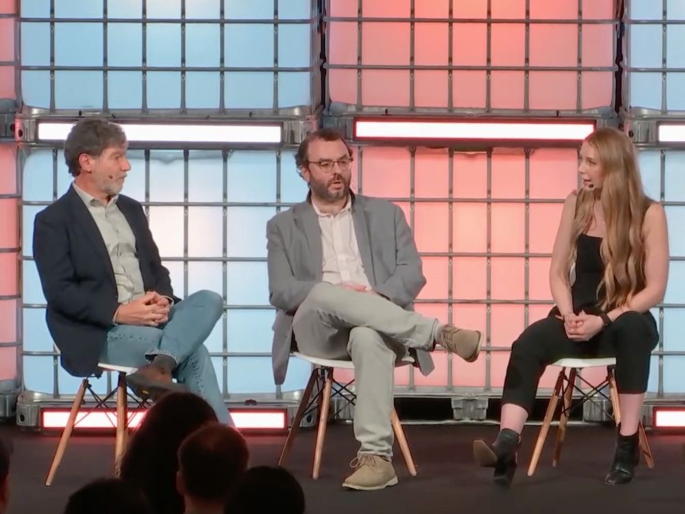 Two men and a woman sit on chairs speaking with one another on a stage with a blue and pink background.