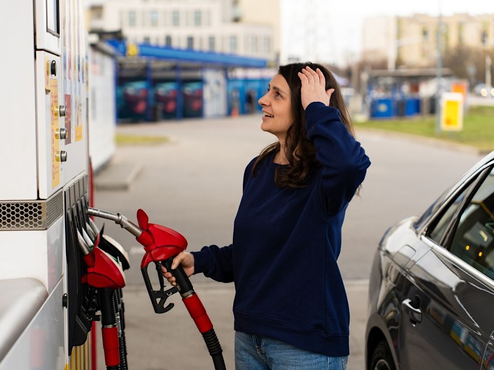 A woman stands at a gas pump, looking at the prices, with one hand on her head.
