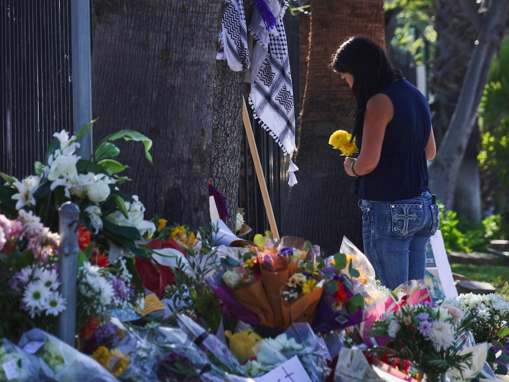 A woman holds yellow flowers. In the foreground, a mound of flowers can be seen against a metal fence. A keffiyeh scarf is also part of the memorial. 