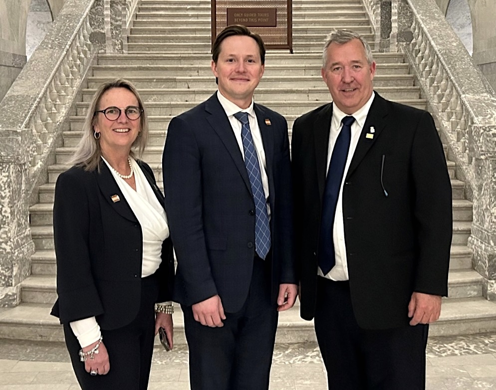 A youngish woman and two men in suits stand in front of a white stone staircase.