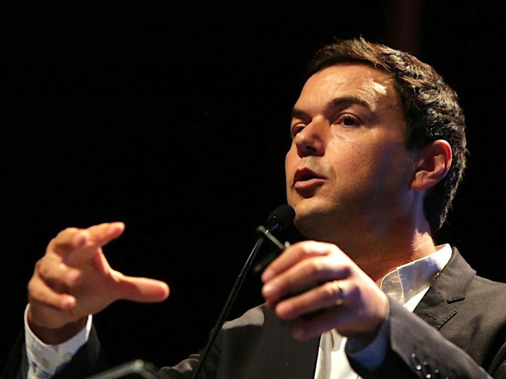 A man with short brown hair and light skin tone speaks into a microphone, gesturing with his hands.