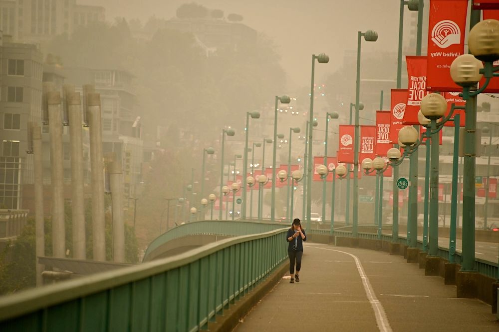 A head-on view of woman in a ball cap reading her mobile phone as she walks across a bridge, the urban background shrouded in brown haze.