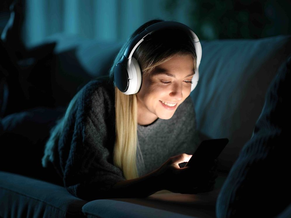 A woman wearing white headphones lies on her stomach on a sofa in a darkened interior space. She is smiling while looking down at her smartphone, its light illuminating her face. She has long blond hair.