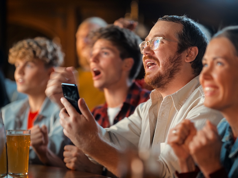 People are at a bar watching a sports game. A man holds a phone.