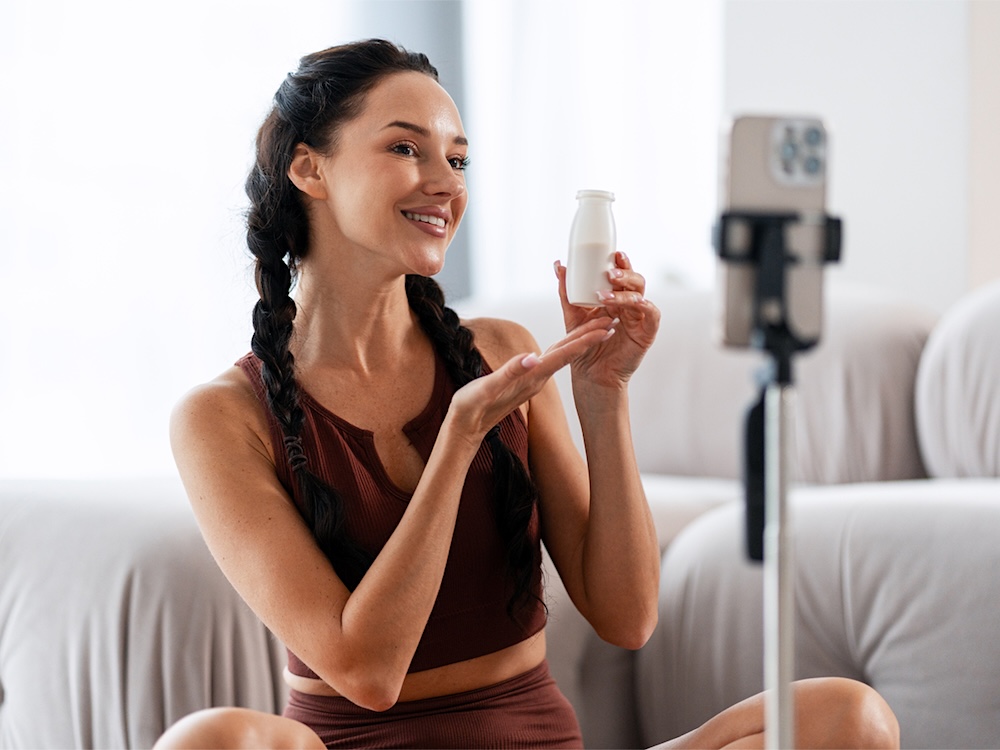 A woman sits in front of a phone on a tripod. She is wearing yoga clothes and holding up a product, presumably to recommend.
