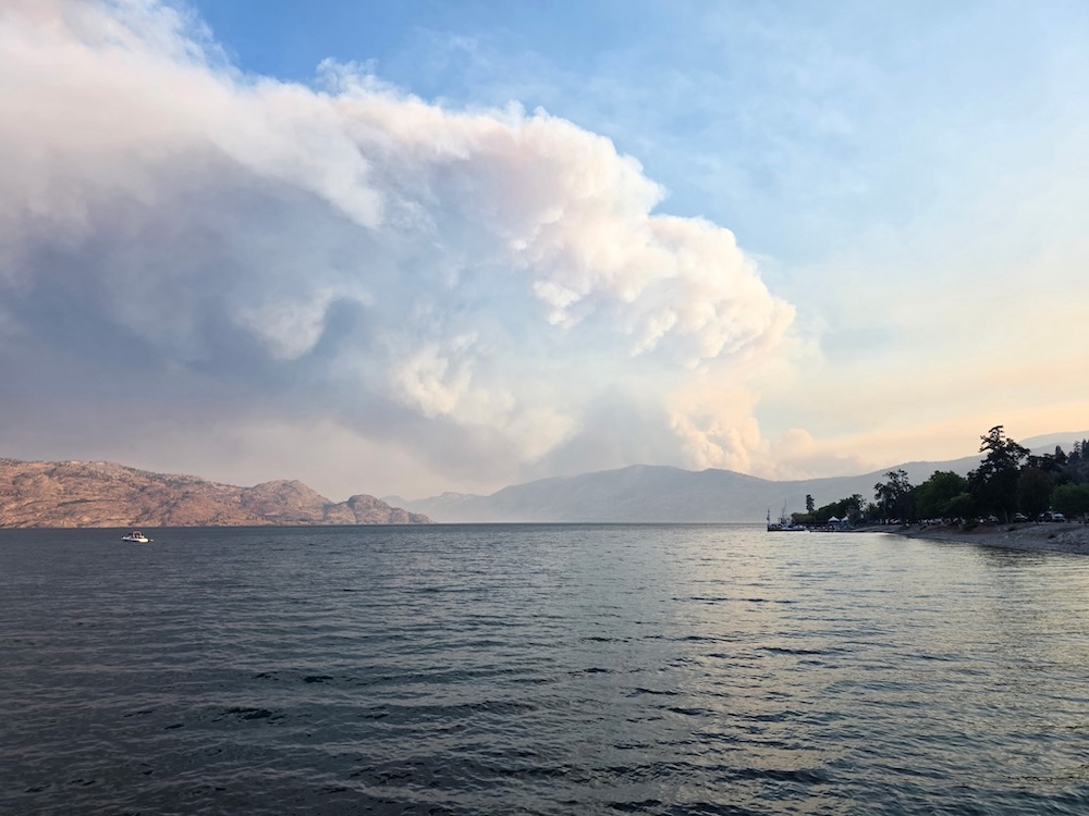 A look across a lake to a distant hill with a giant rising cloud of wildfire smoke.