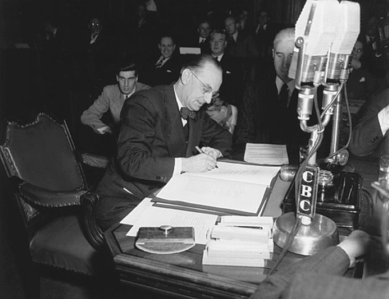 A black and white photo of a light-skinned man with slicked back hair, glasses and wearing a suit with a bow tie. He is seated and signing a large document in a large room with other 