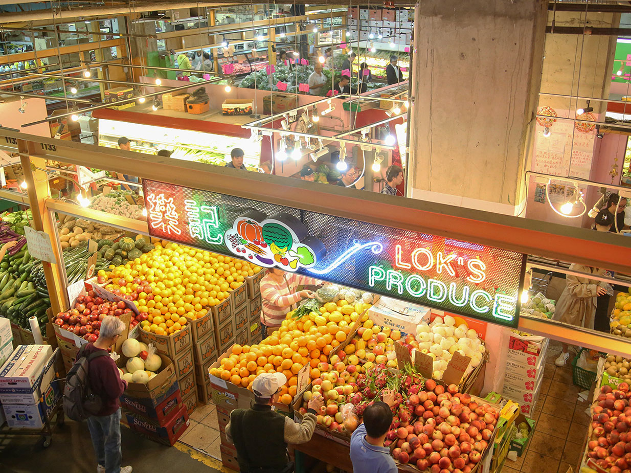 An aerial shot shows customers shopping at Lok&rsquo;s Produce. Fruits and veg are displayed in cardboard boxes, in an abundant indoor farmers market style.
