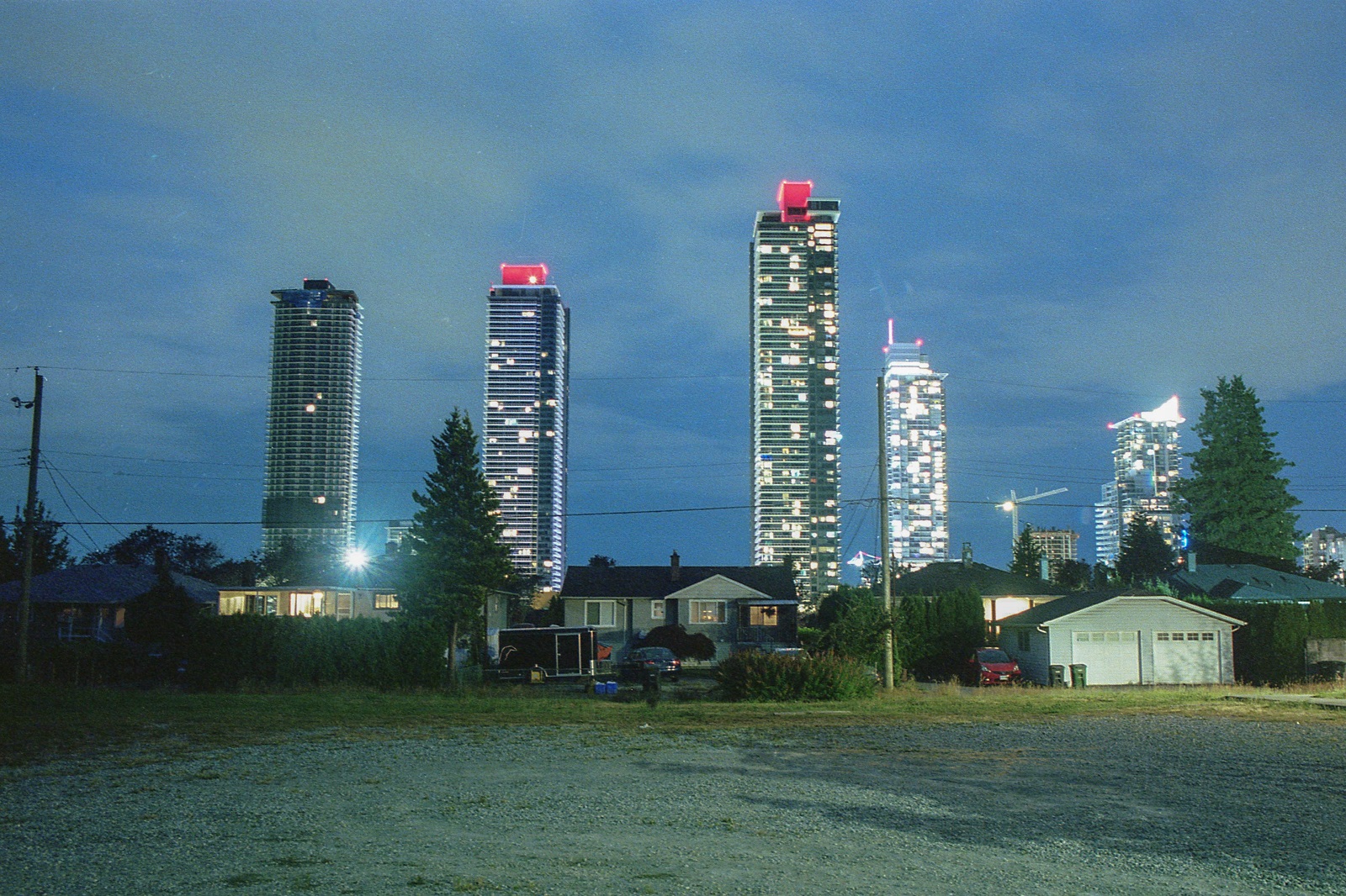 A row of tall towers and construction cranes stand against the darkening sky at dusk. Two towers in the middle have a distinctive topper that glows in red. In front of the towers are older single-family homes that recall a different period of architecture and home building. A wide grass lot is in the foreground.