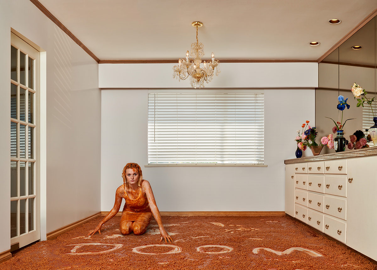 A young woman is on her knees on the floor of a suburban home. To her left is a French door. To her right is a white vanity on which several vases of flowers are arranged in front of the mirror. The woman is wearing a tank dress. Her blond hair and body are covered in baked beans with tomato sauce, which also cover the floor. The word &lsquo;DOOM&rsquo; has been written by sweeping a hand through the beans to expose the white linoleum floor.