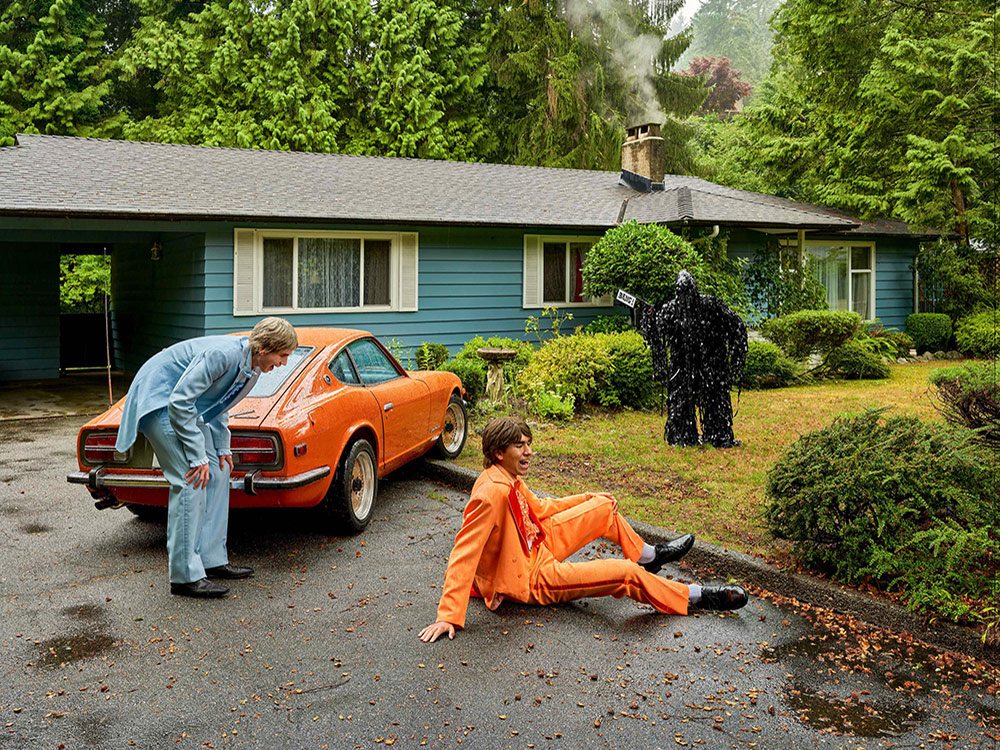 Two young men are wearing bright tuxedos. To the left, a young man with blond hair in light-blue tuxedo is laughing, bending slightly with his hands on his knees. He is standing in front of a bright orange car parked haphazardly over the curb of a driveway that edges onto the front lawn of a suburban bungalow in front of a forest. To the right, his friend has brown hair and is wearing an orange tux the same colour as the car. The young man in orange is seated on the driveway, laughing with his eyes closed. In the front yard, a non-descript monster holds a gun with a sign that reads &ldquo;Bang!&rdquo; The monster seems to be made of unspooled pieces of video tape. 