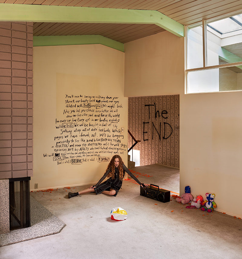 A young woman with long curly brown hair, black lipstick, a black dress and black boots sits in an empty suburban living room. She glares at the camera and rests one arm on a black boombox. The concrete floor is littered with Hawkins Cheezies. To the right, three stuffed animals with blue and pink tape over their faces line the wall. The woman is sitting in front of a handwritten message in Black Sharpie that reads: &lsquo;You&rsquo;ll soon be seeing us walking down your street, our backs held proud, our eyes dilated with truth and power. We might look like you but you should know better. We will draw our line in the sand and force the world to cross our line. Every cell in our bodies explores with the truth. We will be kneeling in front of the Safeway, atop out of date textbooks whose pages we have chewed out. We&rsquo;ll be begging passersby to see the need to question question and question and never stop questioning until the world stops spinning. We&rsquo;ll be adults who smash the tired exhausted system and will crawl and chew our way into a radical new world. We&rsquo;ll change minds and souls from stone + plastic into linen and gold. This is what I believe. This is what I know.&rsquo; Behind her above a staircase, the words &lsquo;THE END&rsquo; are written in black marker.