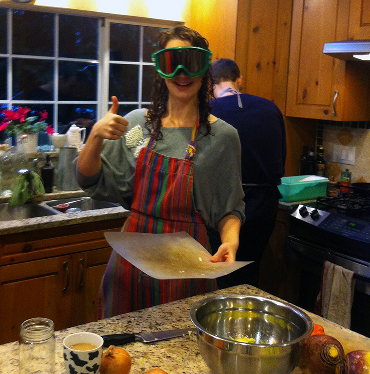 Michelle Gamage, wearing ski goggles and a colourful striped apron, gives the camera the thumbs up. She&rsquo;s holding a plastic cutting board, and there&rsquo;s a metal bowl and some onions on the counter in front of her.