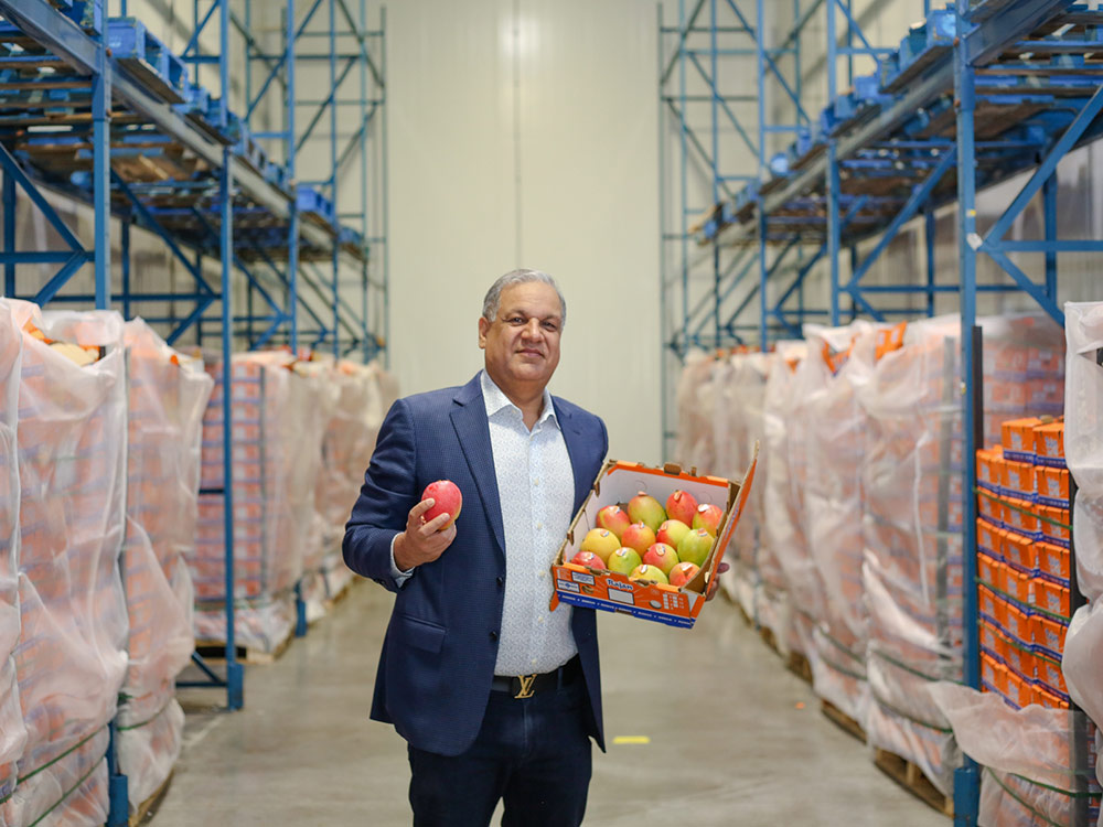Tony Singh wears a blue blazer over a white dress shirt with floral texture. He is standing in the middle of a warehouse. On either side of him are blue shelves holding pallets of Singh&rsquo;s signature Rajan brand Haden mangoes. The orange flats are ensconced in white coverings. In his left arm, Singh is holding an orange box of the mangoes. They have red, orange, yellow and green flesh. Singh holds up one mango for the camera with his right hand. He is smiling.
