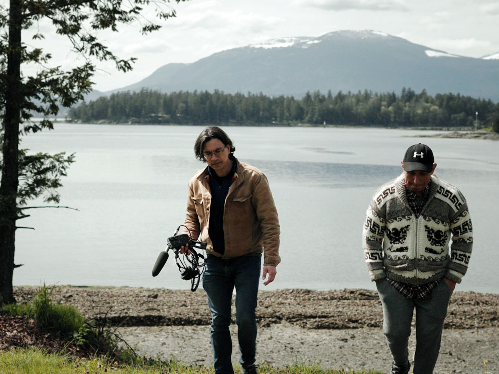 Duncan McCue, left, holding sound recording equipment and a microphone, walks with podcast guest Rocky James on Penelakut. 
