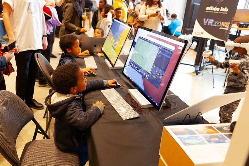 Two kids sit in front of computers. One is playing a game, and the other is looking at an online map.