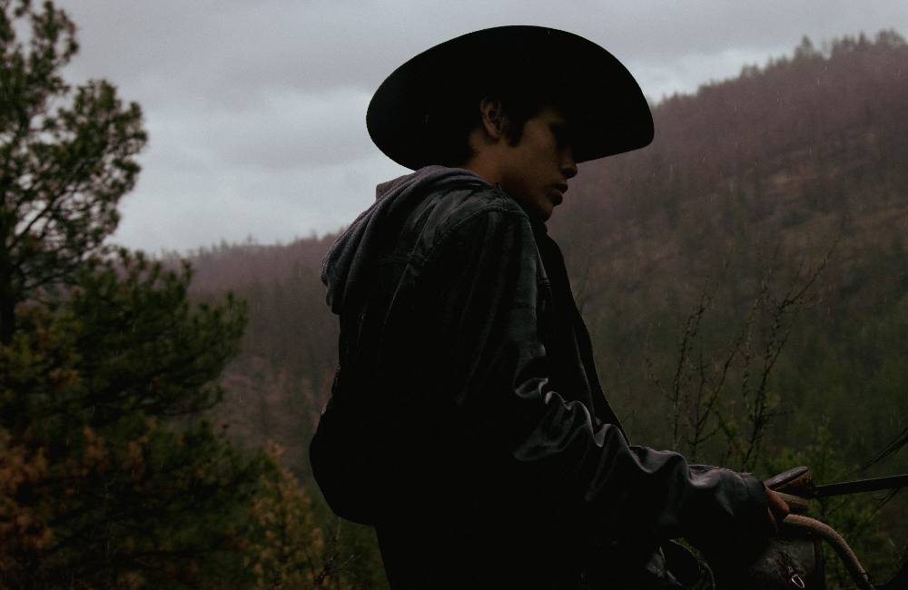 Elijah Swan-Hall sits in the saddle, his black leather jacket slick with rain. He holds the reins in his hands and looks towards the ground.