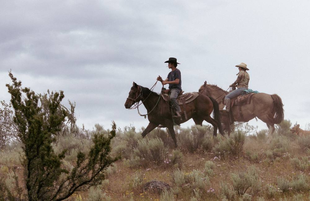 Jessi Wyatt and Elijah Swan-Hall, mounted, walk across the ridge of a hill covered in sagebrush. A cigarette hangs from Swan-Hall&rsquo;s mouth as he lifts the reins.