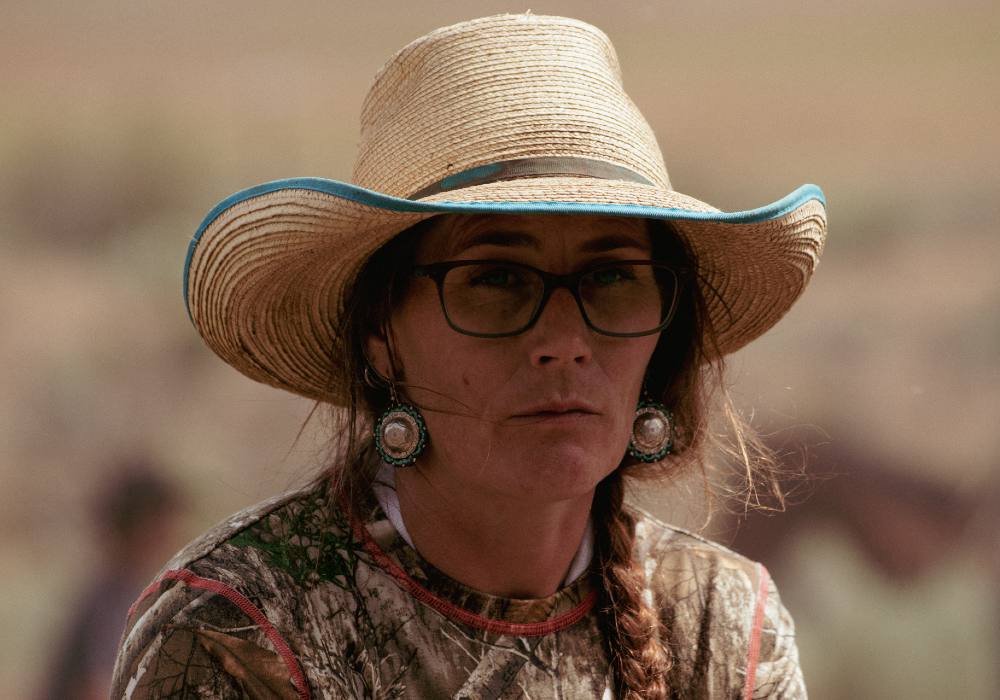 A woman, Jessi Wyatt, looks past the camera. A worn straw hat shadows her face from the sun. She is wearing turquoise and silver medallion earrings. Wisps of hair have escaped her braid and frame her face. 