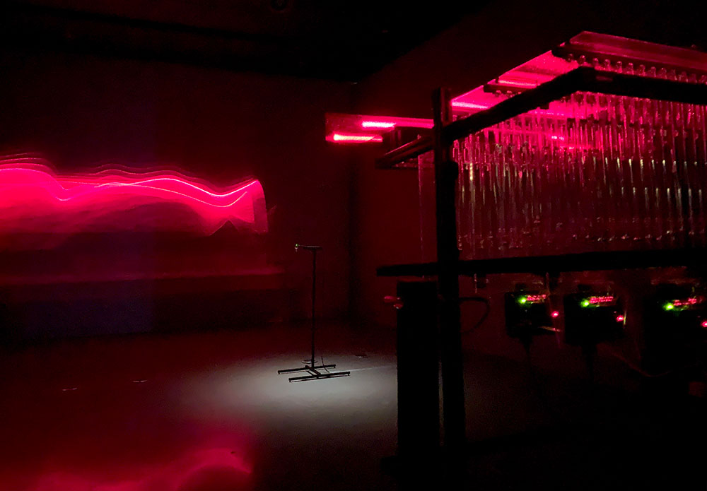 Bright red projections cut through darkness. To the right of the frame, an electronic panel is visible.
The exhibition&rsquo;s introduction places the individual pieces in the show into this human context: &ldquo;Ancient cosmology was rooted in our meticulous observation of natural patterns. Hundreds of thousands of years in the making, the most sensitive, sophisticated, and flexible instrument of observation may still be us; our frail bodies and creative minds.&rdquo; 