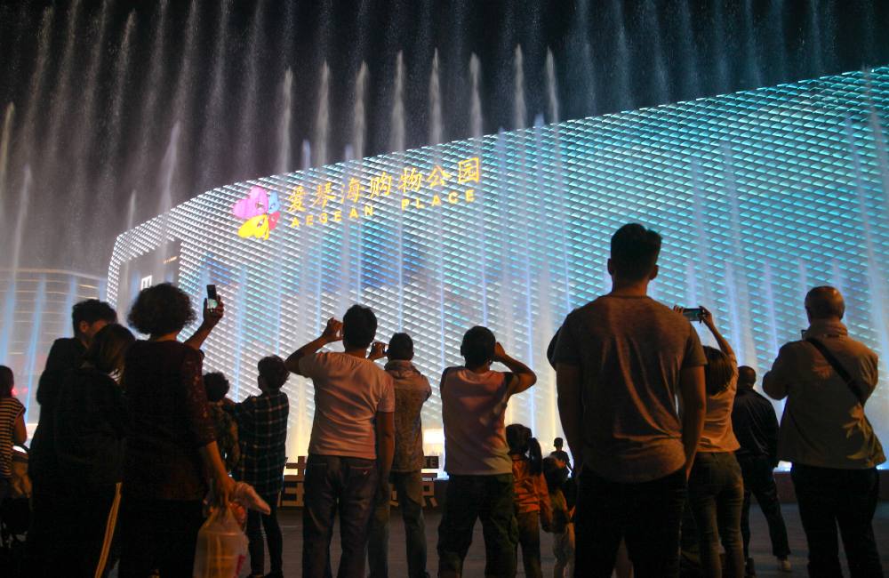 Silhouettes of people photograph a fountain show in front of giant glowing mall at night.