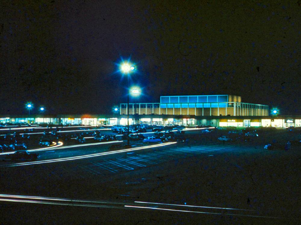 It&rsquo;s night and a boxy department store rises behind a strip of connected shops, all aglow in the dark. In the foreground is a large parking lot with a smattering of cars. The scene is lit with a few tall lamps.