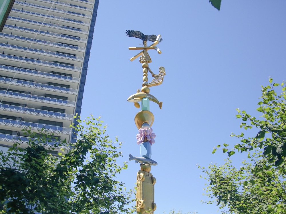 A tall, pastel coloured art installation stands against a bright blue sky. The work, by local artist Douglas Coupland, is titled Charm Bracelet and brings together ephemera that are said to represent the interplay between Burnaby&rsquo;s culture and its retail and civic spaces.