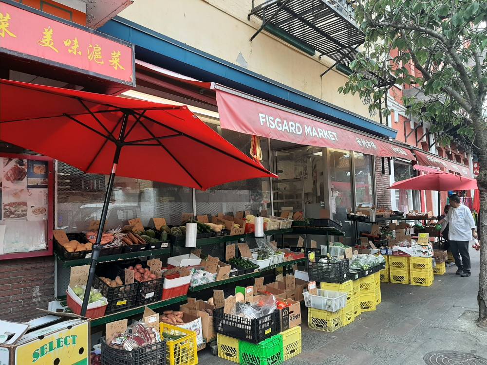 A sidewalk view of Victoria&rsquo;s Fisgard Market shows cases of produce stacked on outdoor shelves under bright red awnings.