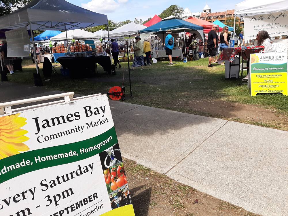 The entrance to James Bay Market, a farmers market in Victoria, features a colourful sandwich board that reads "James Bay Community Market.&rdquo; There are several tented vending stalls in the background.