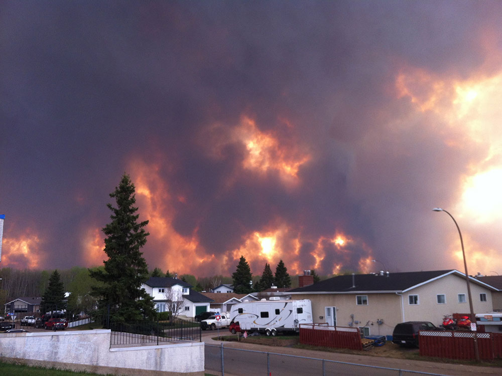 Suburban houses are in the foreground with a terrifying wall of flame and grey smoke behind them.