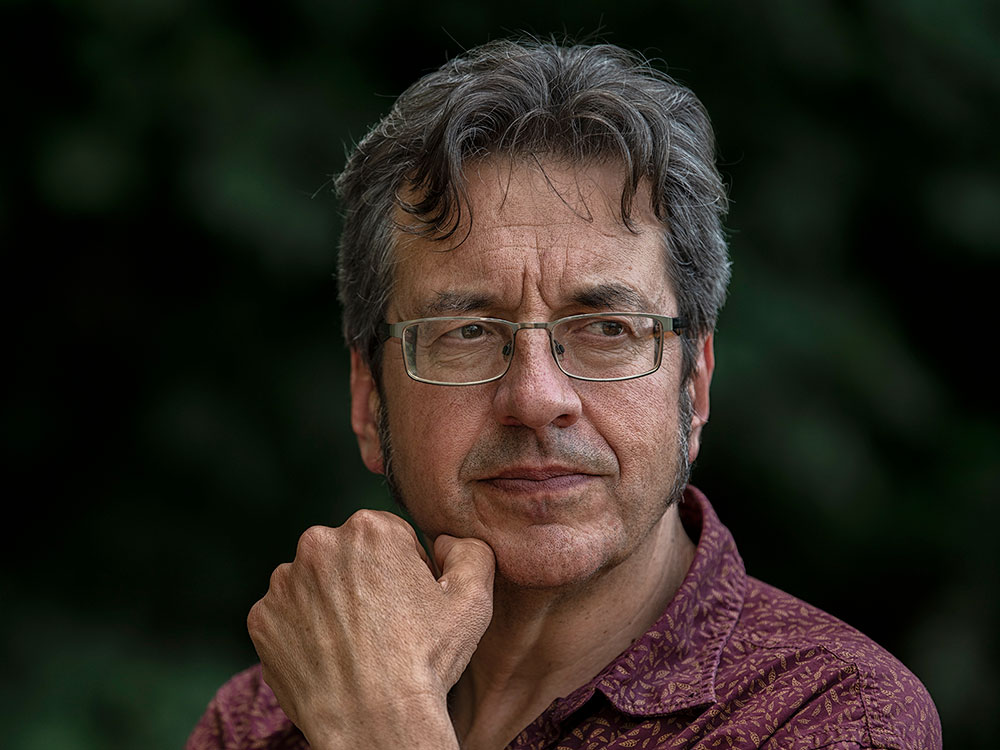 A man wearing glasses stands outside. He has brought his hand up to his chin in a pensive gesture.
