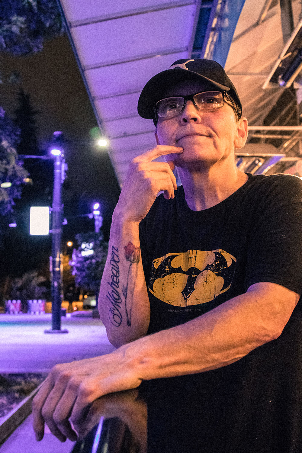 A middle-aged woman leans against the bannister of a patio. On her forearm is a large tattoo of a rose with the words “Maheaven” framing it.