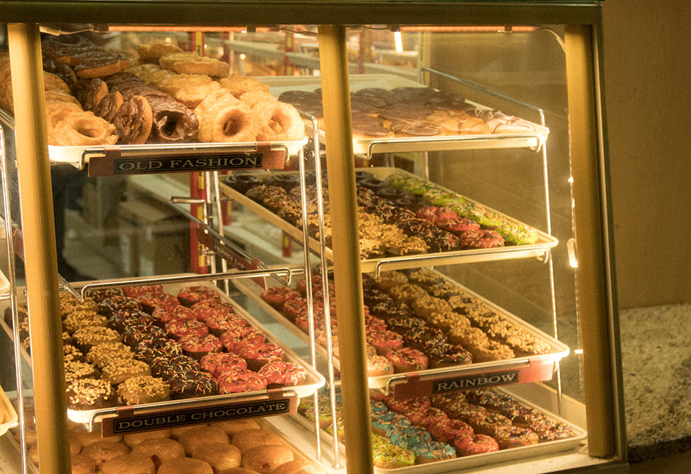Seven trays of doughnuts are displayed behind a glass cabinet. Flavours include ‘old fashioned,’ ‘rainbow’ and ‘double chocolate’. The glazed sugar and sprinkles glistens in the soft lights.