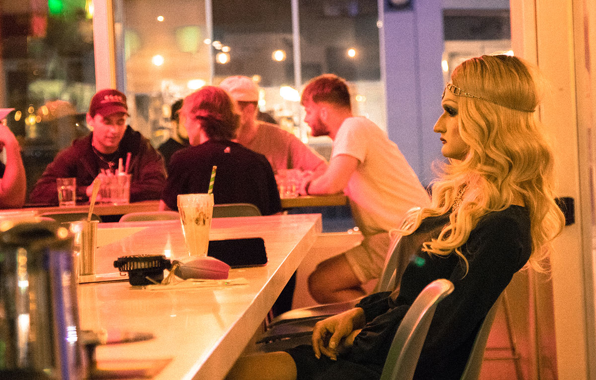 Glimmer, a drag queen with flowing blonde hair, sits at the counter. Her hands are folded in her lap as she stars into space, a half-finished chocolate milkshake in front of her.