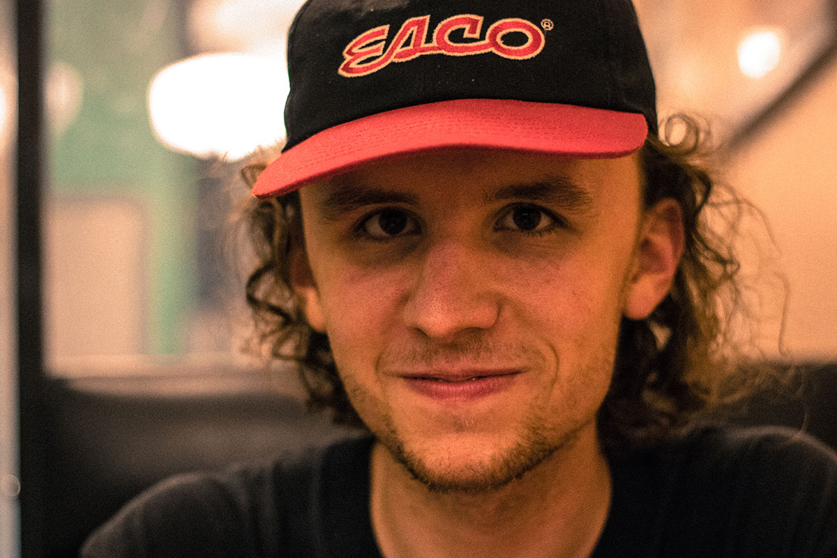 A man, early-to-mid-20s with curly hair tucked under a black and red ball cap, smiles at the camera.
