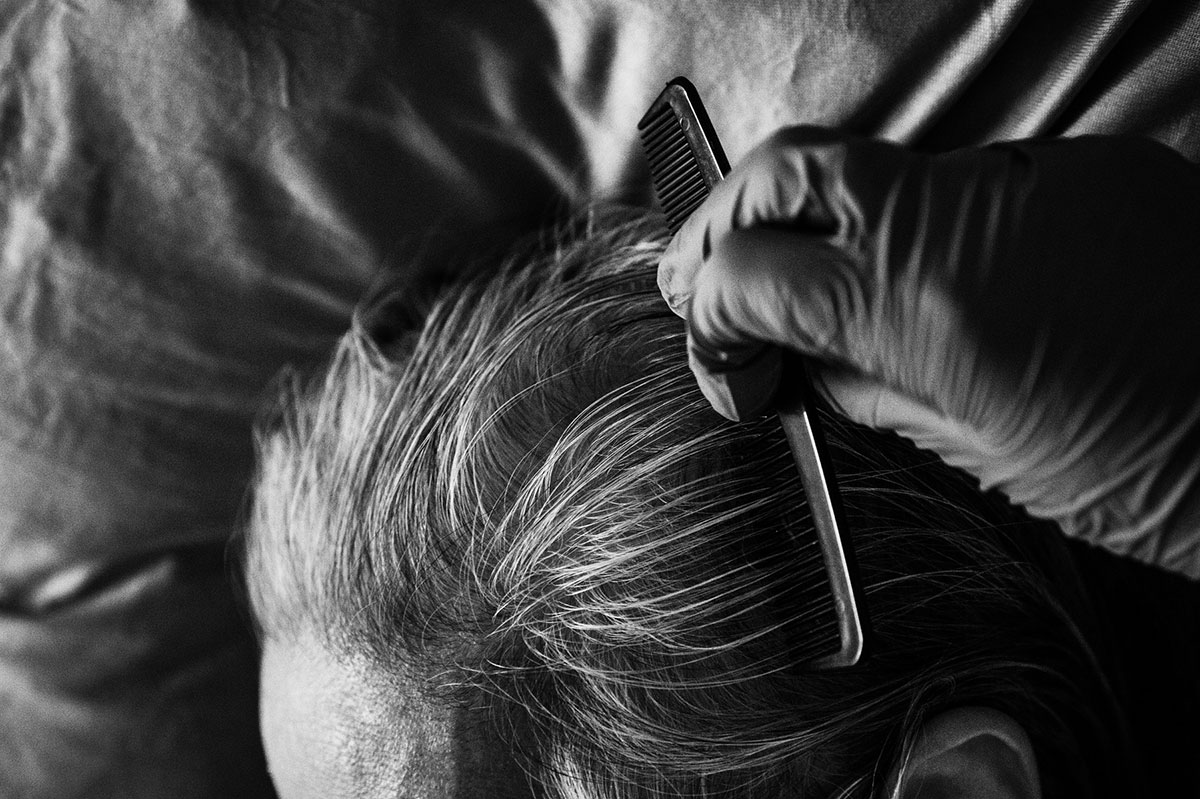 A close up of an elderly woman&rsquo;s hair, with a hand wearing a medical glove running a comb through it.