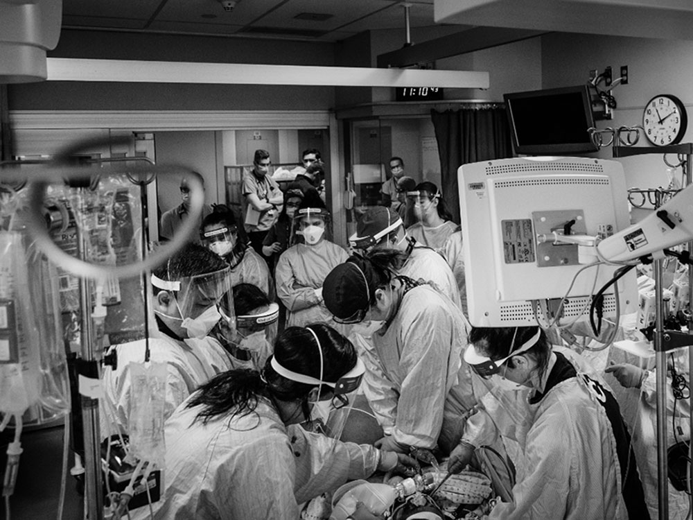 Doctors and nurses crowd around a patient&rsquo;s bed, all wearing masks and face shields.