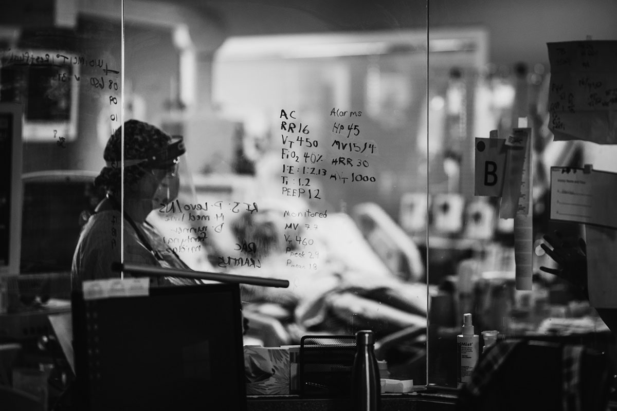 A nurse wearing a mask and face shield in a hospital&rsquo;s isolation room, seen through a window with ventilator settings written on its surface.