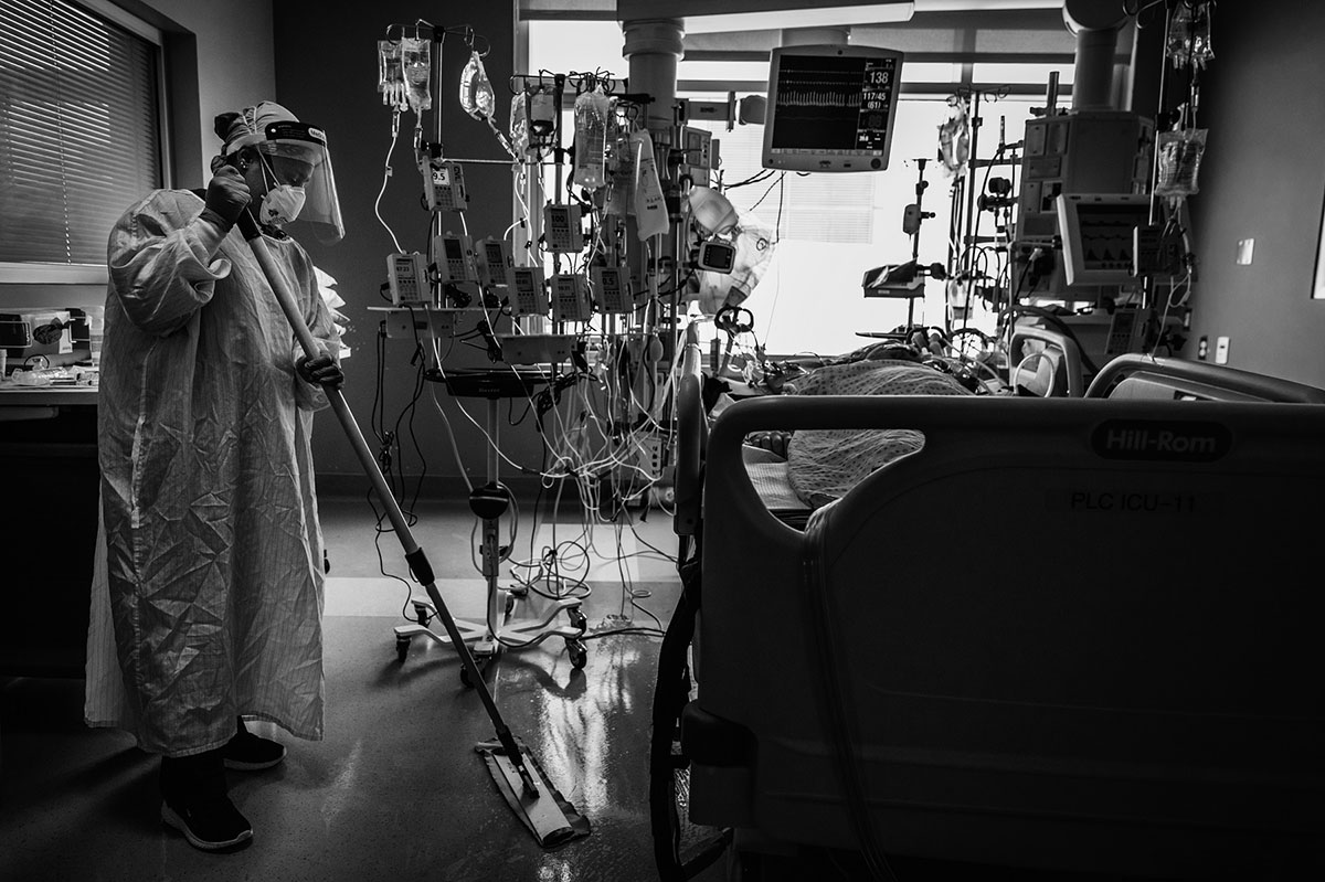 A woman in full protective equipment &mdash; her mask, her face shield, her gown &mdash; mops a hospital room. There is a patient in the bed to the right and over a dozen IV drips next to him.