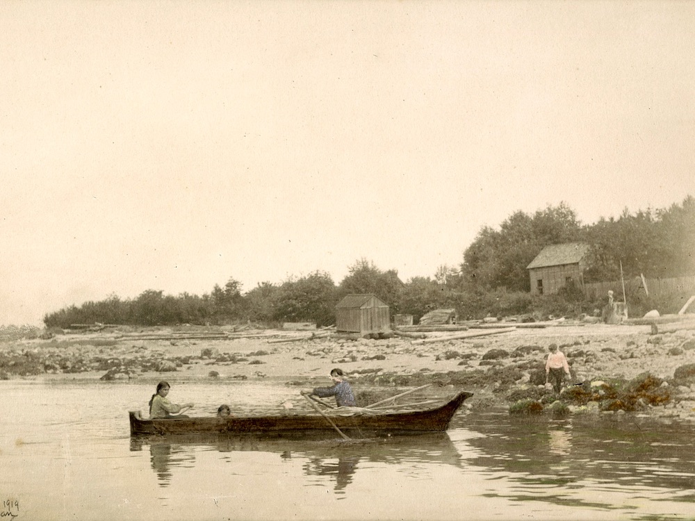 An archival photo from circa 1907 depicts the shores of the Kitsilano Indian Reserve looking east. A man in a blue shirt, Sḵwx̱wú7mesh Chief August Jack Khatsahlano, pulls the oars on a dugout canoe. His wife Swanamia (Marrian) is seated in the canoe to the left of the frame, and a child is in the middle of the boat. Another child is walking along the rocky shoreline nearby. The silver gelatin print has some areas hand drawn in by coloured pencil.
